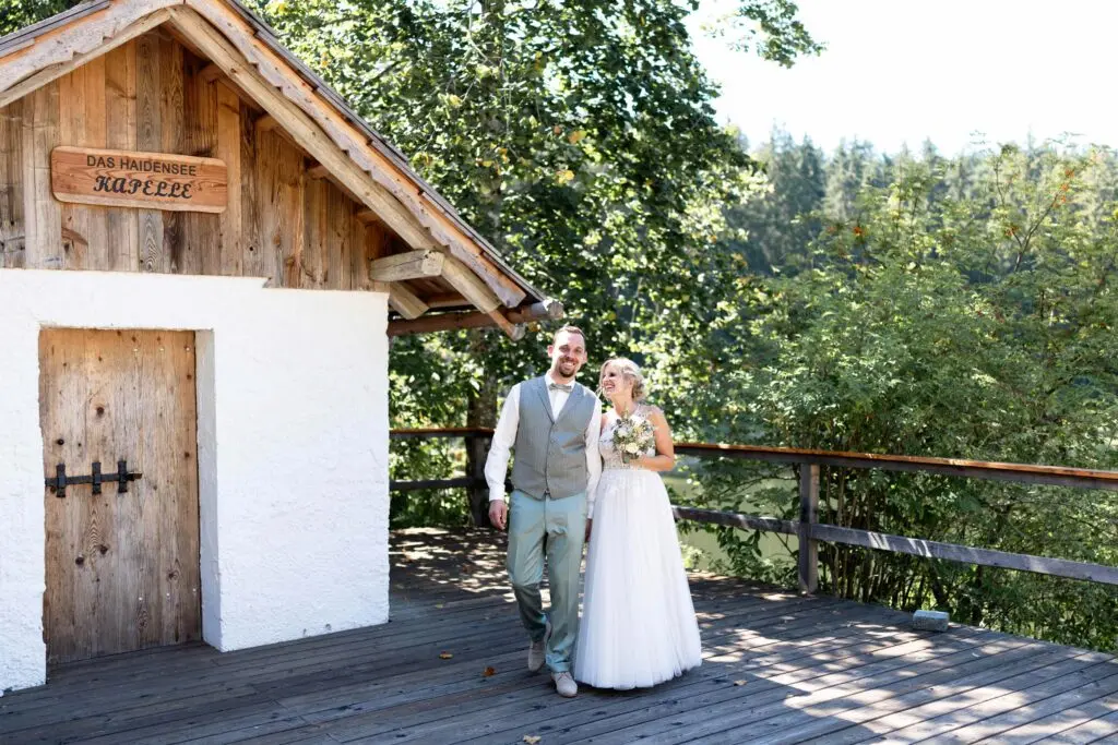 Brautpaar in Weiß und Anzug geht Hand in Hand über den Steg am dasHaidensee mit Kapelle im Hintergrund