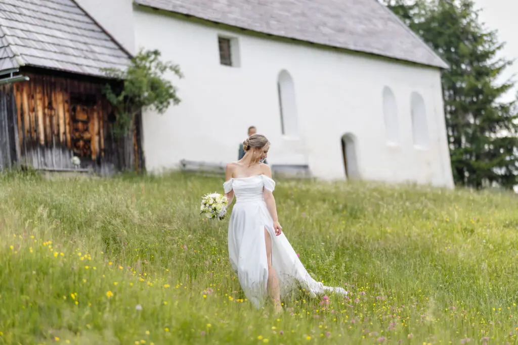 Braut im weißen Hochzeitskleid auf einer Almwiese vor einer Kirche auf der Turracher Höhe