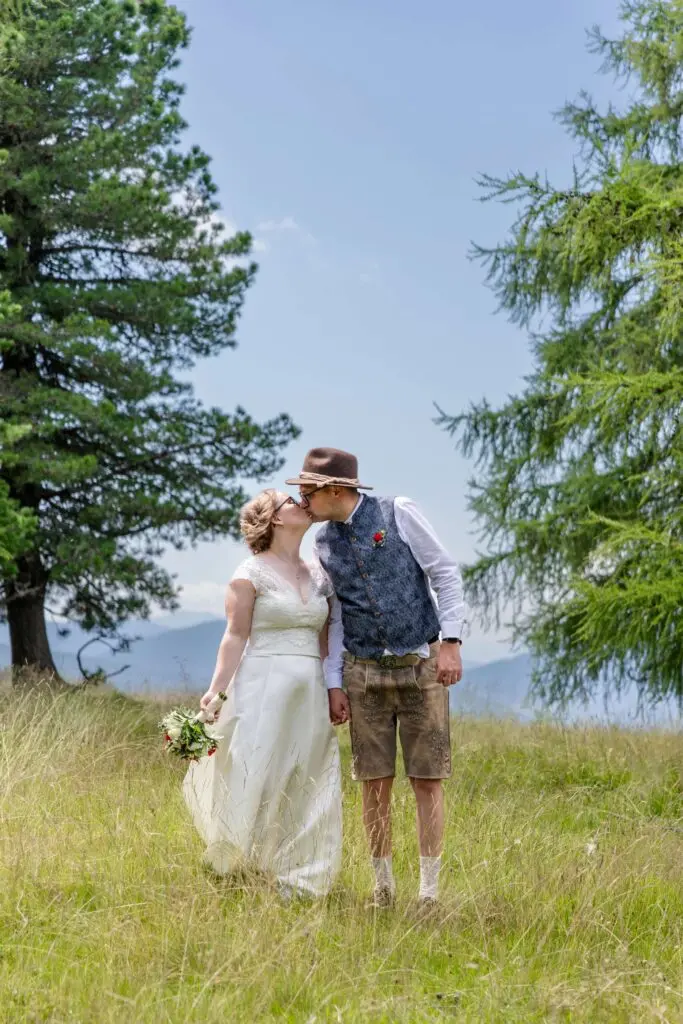 Braut im weißen Hochzeitskleid und Bräutigam in Tracht küssen sich auf der Hochrindl Alm